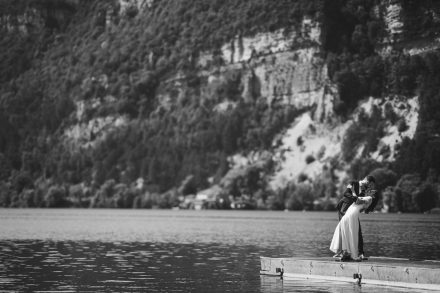 Photographie en noir et blanc de deux élégantes mariées enlacées sur un ponton au bord d'un lac d'altitude, capturée avec un objectif 200mm
